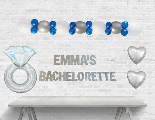 Decorative setup for a bachelorette party with balloons and 'Emma's Bachelorette' text on a white brick wall.