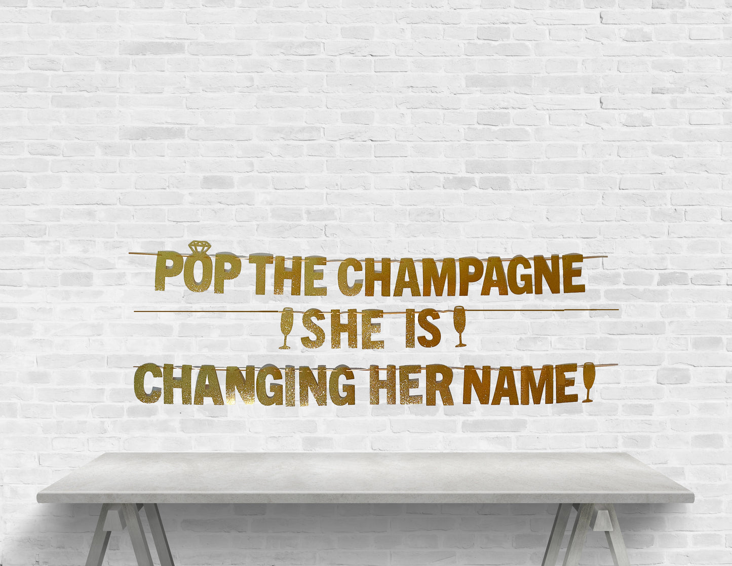 Text 'Pop the Champagne! She is changing her name!' on a white brick wall with a table in front.